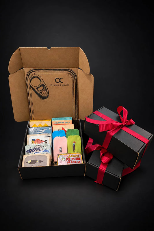 Open Oysters & Caviar tinned fish mystery box with assorted tins, beside two black gift boxes with red ribbon